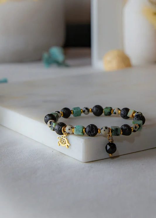 Afrykańska Harmonia Bransoletka Z Kamieniami z turkusowymi i czarnymi kamieniami, złotym żółwikiem i zawieszką na marmurowej podstawie.