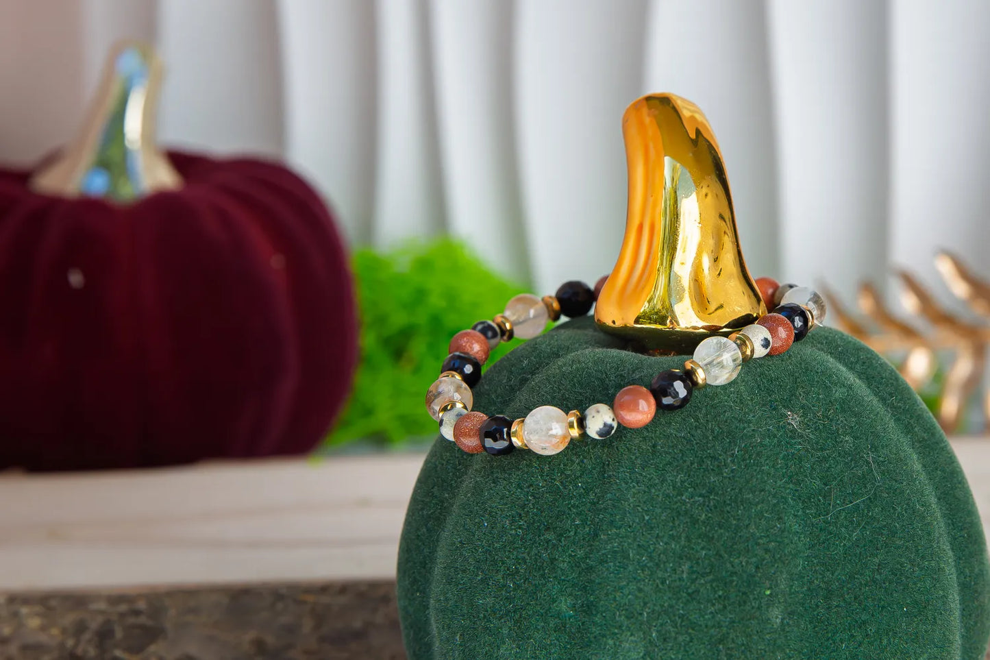 ALCHEMIA SUKCESU - BRANSOLETKA Z CYTRYNU, PIASKU PUSTYNI I KRYSZTAŁU GÓRSKIEGO. Bransoletka z kolorowymi kamieniami, w tym cytrynami, piaskiem pustyni i kryształem górskim, umieszczona na zielonym dyniowym podstawce. Tło z dyni i liści.