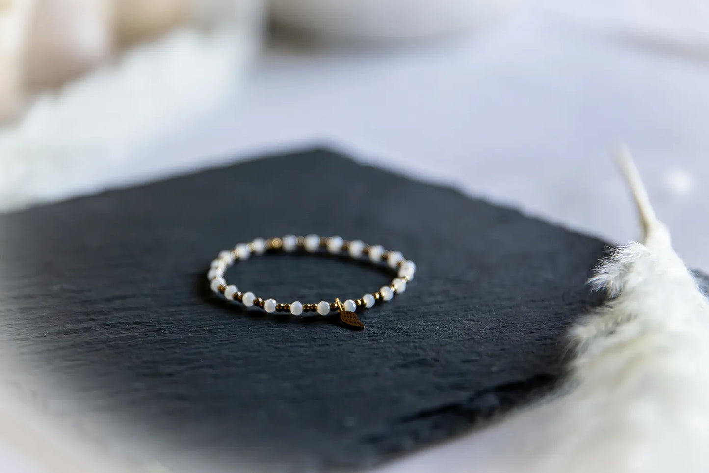 Anielski Blask Bransoletka Z Hematytu I Porcelany Delikatna, z białymi kamieniami i złotymi hematytami, na ciemnej płycie, z piórkiem w tle.