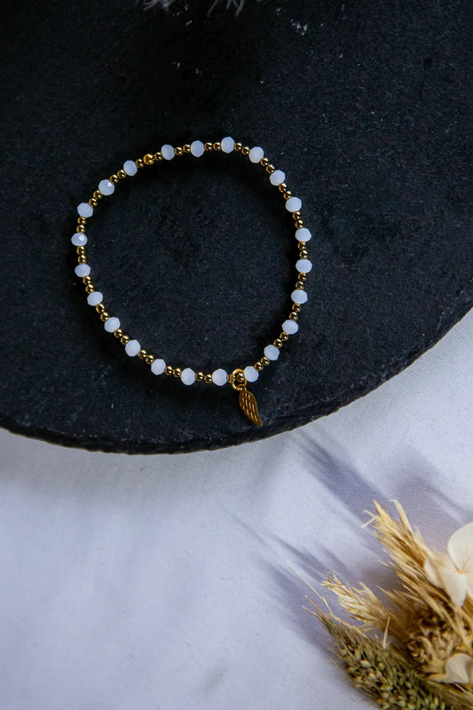 Anielski Blask Bransoletka Z Hematytu I Porcelany Elegancka z złotym zawieszeniem w kształcie skrzydła na czarnym tle.