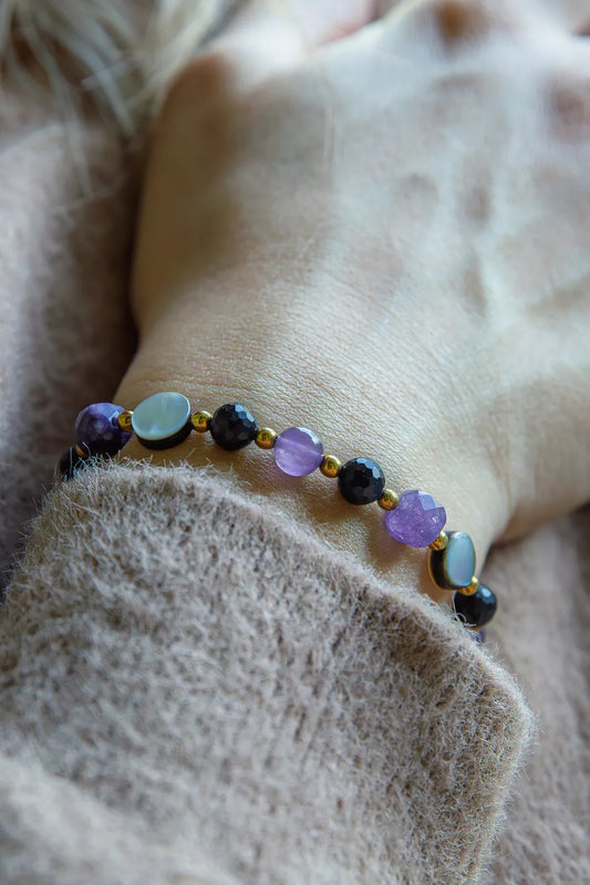 BLASK KSIĘŻYCA - BRANSOLETKA Z AMETYSTU, MASY PERŁOWEJ I CZARNEGO TURMALINU. Bransoletka z kolorowymi kamieniami, w tym ametystem, masą perłową i czarnym turmalinem, na tle delikatnej, beżowej tkaniny.
