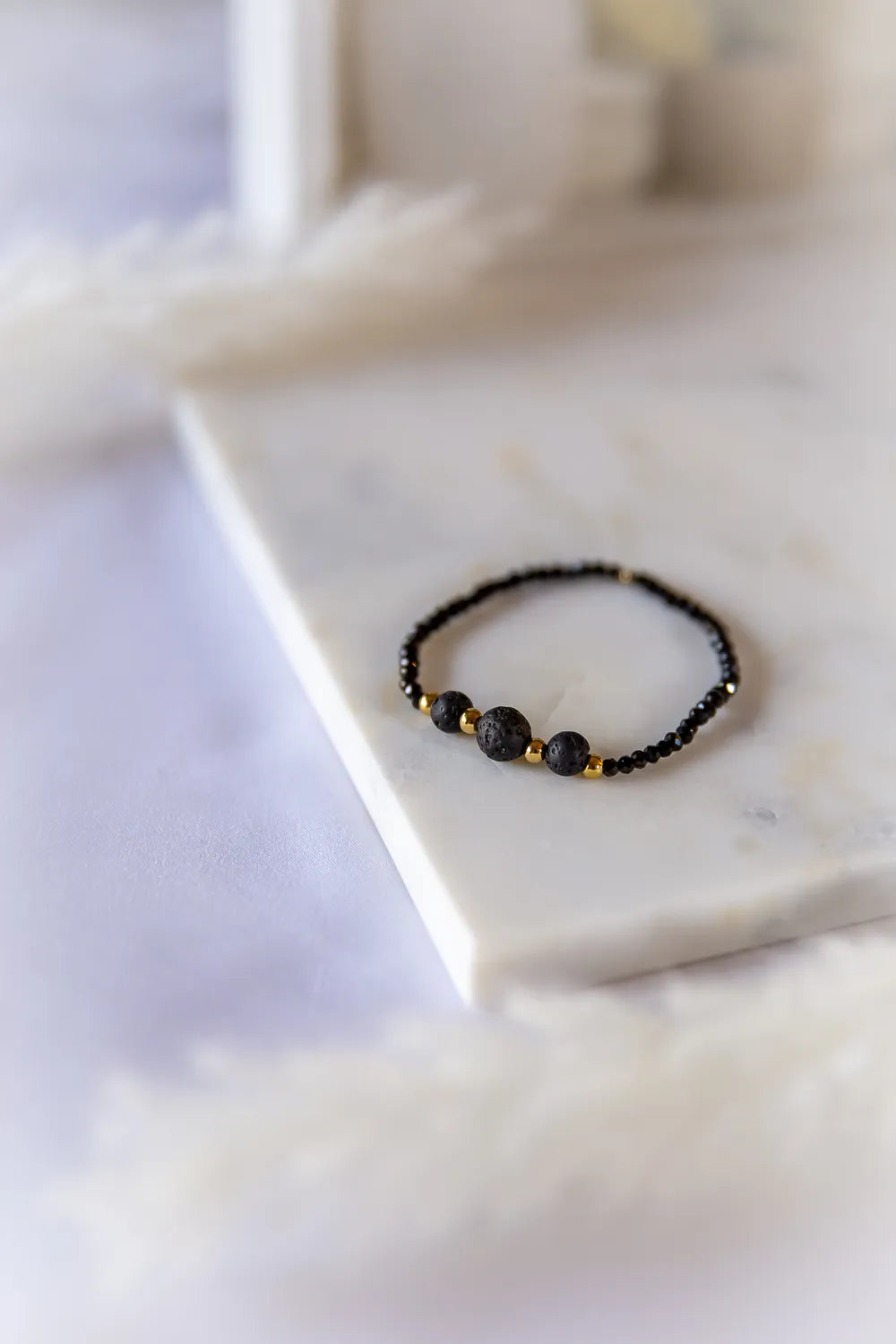 Czarna Moc Bransoletka Czarne Kamienie: subtelna, lecz wyrazista, z okrągłymi czarnymi kamieniami lawowymi i złotymi przekładkami na tle marmuru.