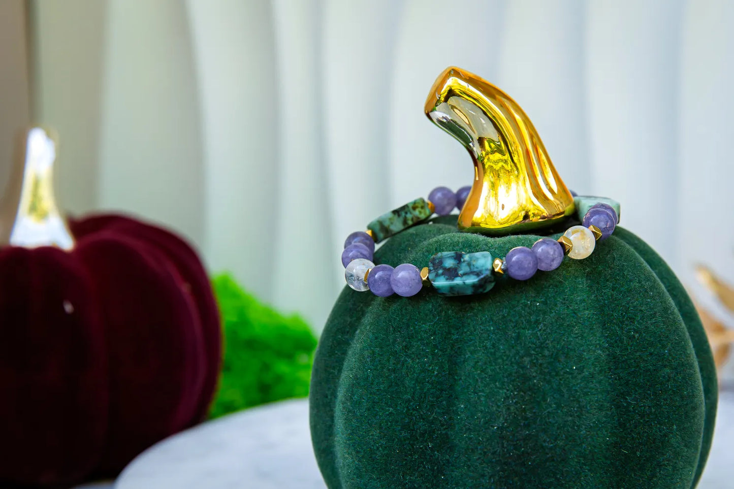 DROGA DUSZY - BRANSOLETKA Z TURKUSU AFRYKAŃSKIEGO, AMETYSTU I CYTRYNU. Bransoletka z kolorowymi kamieniami, w tym turkus, ametyst i cytryn, umieszczona na zielonym aksamitnym dyniowym podstawce. Tło z czerwonymi i zielonymi dekoracjami.