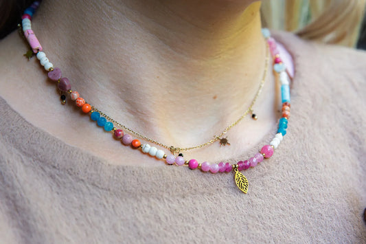 FESTIWAL RADOŚCI - NASZYJNIK Z JADEITU TURMALINOWEGO, TURMALINU I JASPISU. Naszyjnik z kolorowymi kamieniami, w tym jasnopinkowymi, pomarańczowymi i niebieskimi, na złotym łańcuszku. Tło przedstawia delikatną skórę i beżowy sweter.