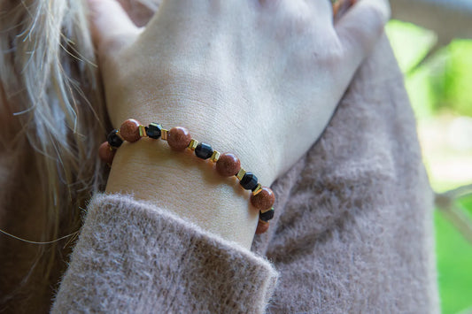 GWIEZDNA PUSTYNIA - BRANSOLETKA Z PIASKU PUSTYNI I CZARNEGO AGATU na nadgarstku kobiety. Biżuteria składa się z okrągłych kamieni piasku pustyni i czarnego agatu, ułożonych na tle delikatnej, beżowej bluzy.