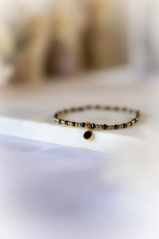 Klasyczna Elegancja Bransoletka Hematyt Turmalin z czarnymi, błyszczącymi kamieniami i złotymi koralikami, ozdobiona owalnym czarnym wisior. Delikatne ułożenie na białym tle.