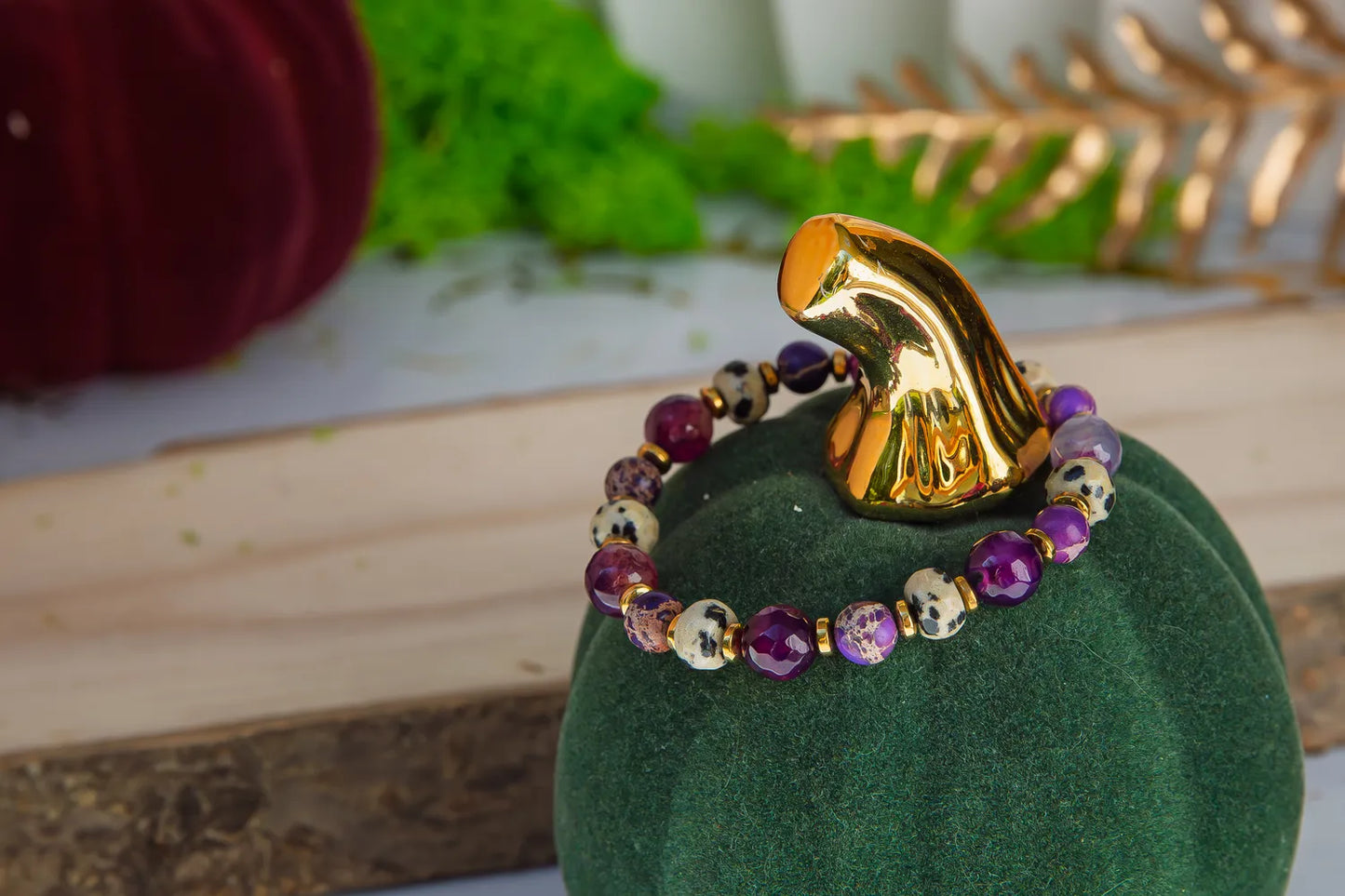 KRÓLEWSKA INTUICJA - BRANSOLETKA Z JASPISU CESARSKIEGO I FIOLETOWEGO AGATU. Bransoletka z kolorowymi kamieniami, w tym jaspisem cesarskim i fioletowym agatem, na zielonym aksamitnym podstawce. Tło z dekoracjami roślinnymi i złotymi elementami.