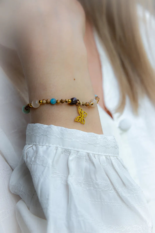 Motyli Taniec Bransoletka Z Kamieniami: złoty motyl zawieszka na nadgarstku, z koralikami: niebieskimi, białymi, brązowo-złotymi i fioletowymi. W tle jasna, delikatna tkanina.