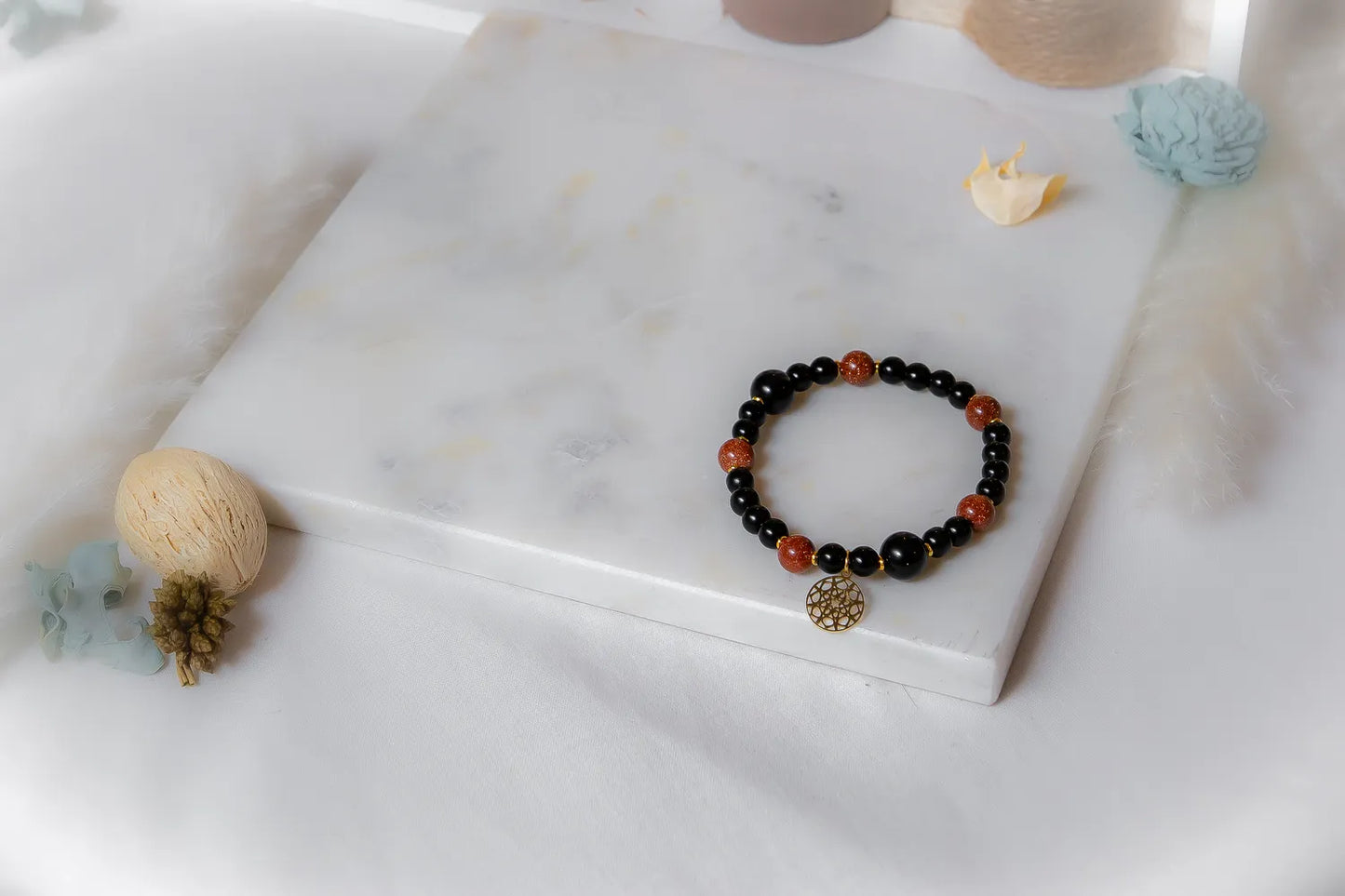 Pustynna Moc Bransoletka Xl Piasku Pustyni Czarny Onyks: czarna bransoletka z koralików z kamieniami piasku pustyni i okrągłą złotą zawieszką, ułożona na białej marmurowej płycie.