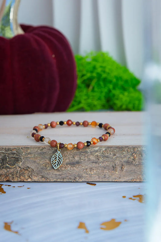 SERCE JESIENI - BRANSOLETKA Z KARNEOLU, GRANATU I CYTRYNU, złożona z kolorowych kamieni i zawieszką w kształcie liścia, leżąca na drewnianym tle z czerwoną dynią i zielonym mchem w tle.