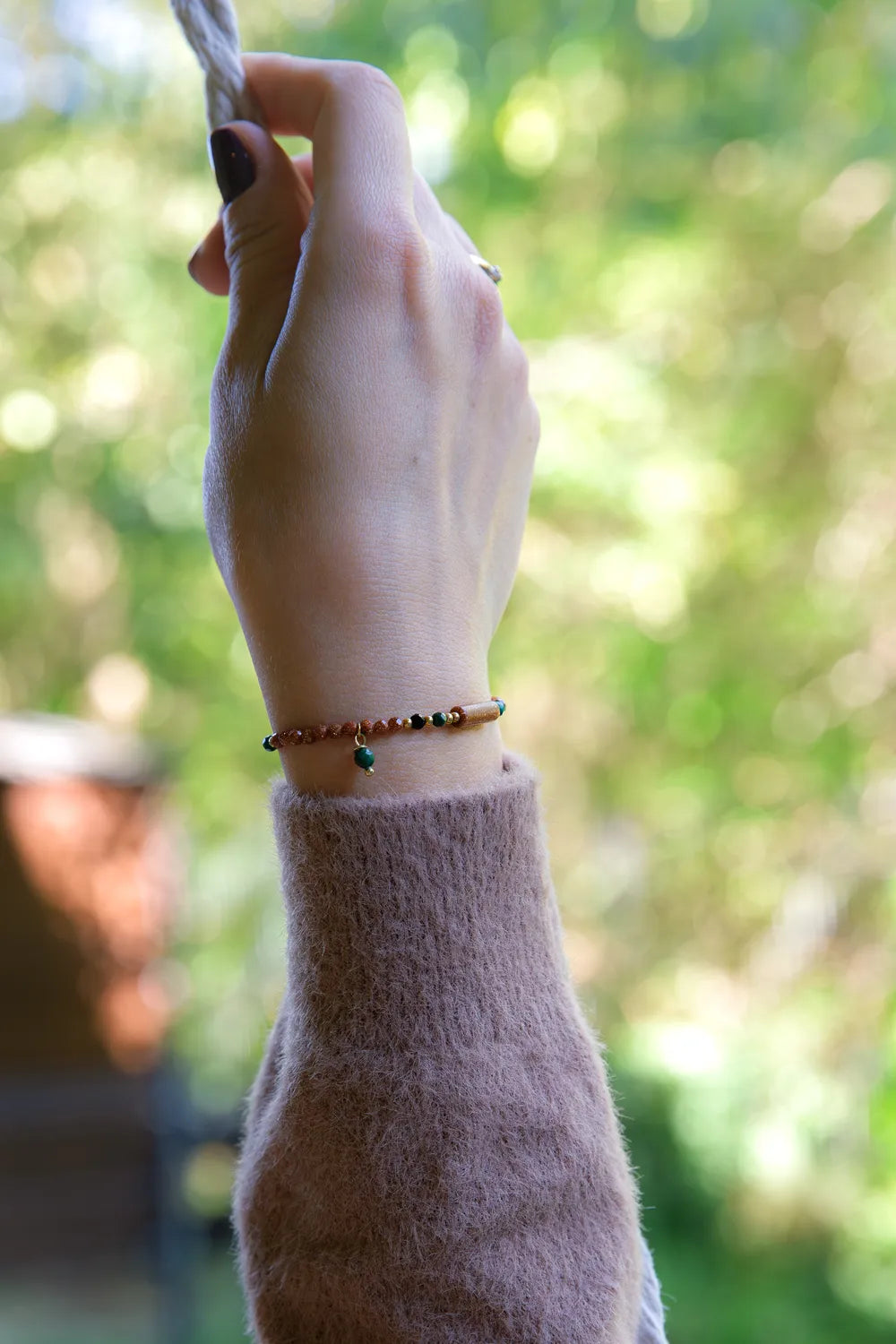 SZEPT LASU - BRANSOLETKA Z PIASKU PUSTYNI, AGATU I TURMALINU na nadgarstku, widoczne koraliki z piasku pustyni, agatu i turmalinu, tło z zielonymi plamkami światła.