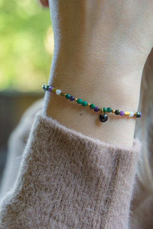 TAJEMNY OGRÓD - BRANSOLETKA Z LEPIDOLITU I CHALCEDONU na nadgarstku, z kolorowymi kamieniami i czarnym zwisem, na tle zielonego tła.