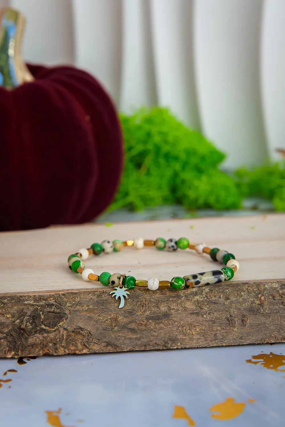 TWOJA OAZA - BRANSOLETKA Z ZIELONEGO JASPISU CESARSKIEGO I HOWLITU. Bransoletka z kolorowymi koralikami zielonego jaspisu cesarskiego i howlitu, ułożona na drewnianym tle z zielonymi roślinami w tle.