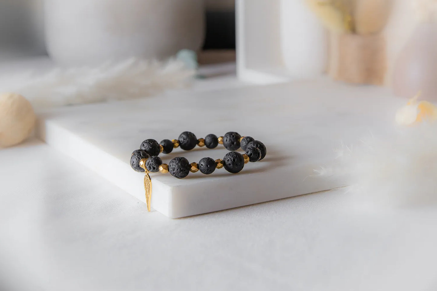 Złote Skrzydlo Bransoletka Czarne Kamienie z złotą zawieszką skrzydła, na białej marmurowej powierzchni, delikatne tło z piórami.