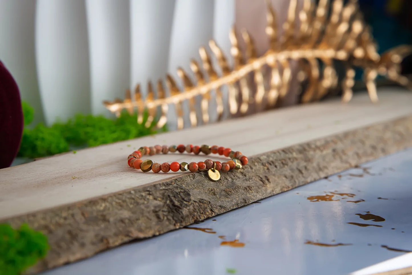 ZŁOTO PAŹDZIERNIKA - BRANSOLETKA Z JASPISU CESARSKIEGO I HEMATYTU, prezentująca się na drewnianym tle z zielonymi dodatkami. Biżuteria składa się z kolorowych kamieni i małych złotych elementów.