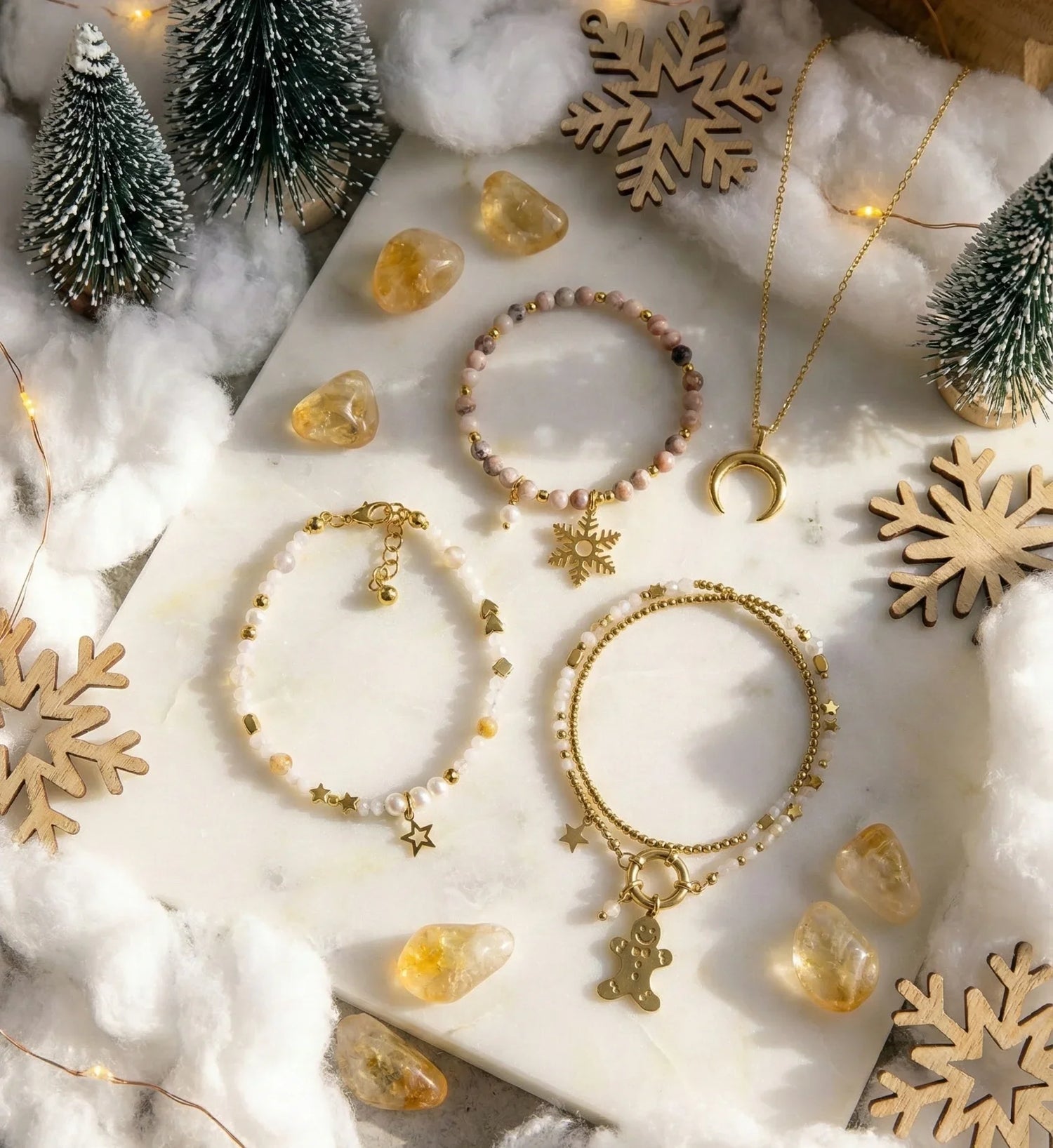Zimowa biżuteria Pati Linka: złote bransoletki z kamieniami naturalnymi i zawieszkami (śnieżynka, gwiazdki, piernik) oraz naszyjnik z księżycem. Świąteczna kompozycja z cytrynem i śniegiem, idealna na prezent.