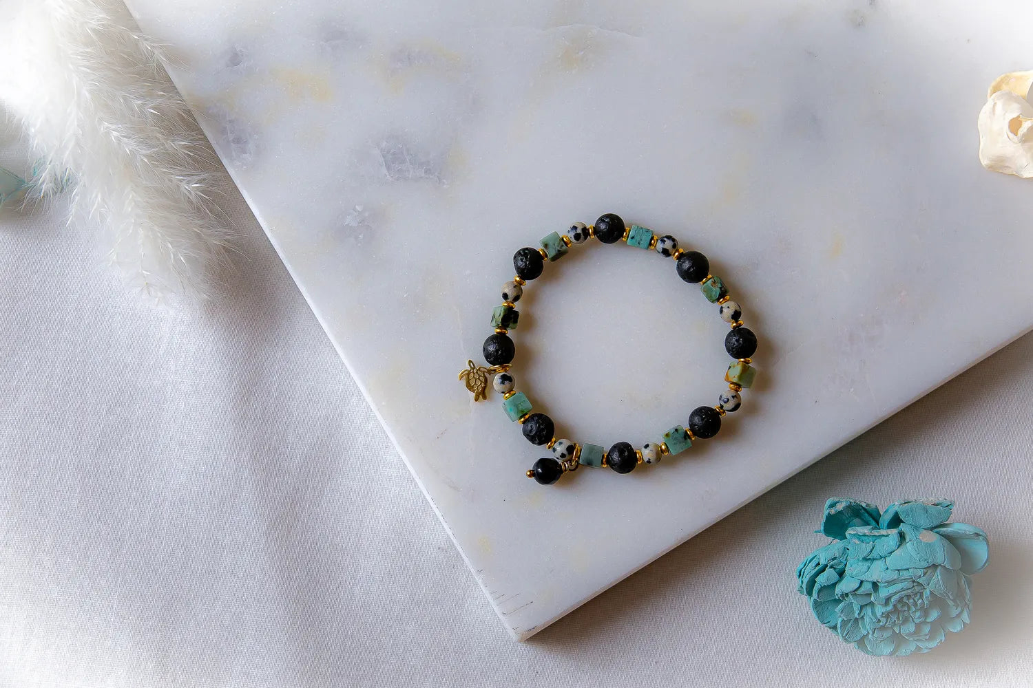 Afrykanska Harmonia Bransoletka Kamienie: okrągłe, czarne kamienie, kostki jaspisu i kamienie w cętki, ze złotą zawieszką żółw, na marmurowym tle.