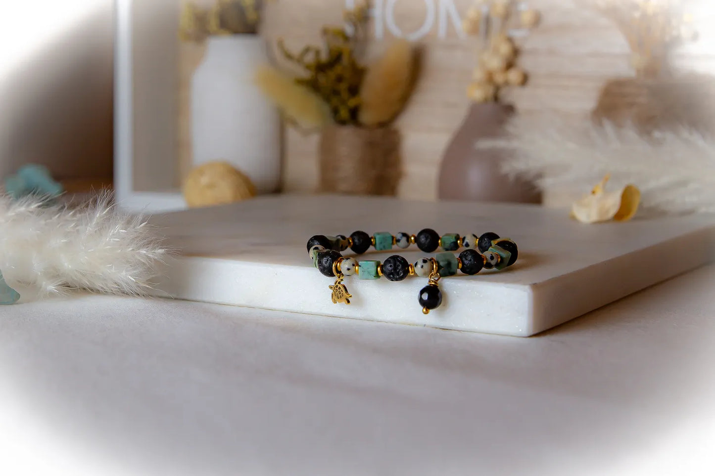 Afrykanska Harmonia Bransoletka Z Kamieniami: bransoletka z kamieni czarnego lawy, jaspisu i turkusowych kwadratów, z złotymi zawieszkami, na białym tle z dekoracjami.