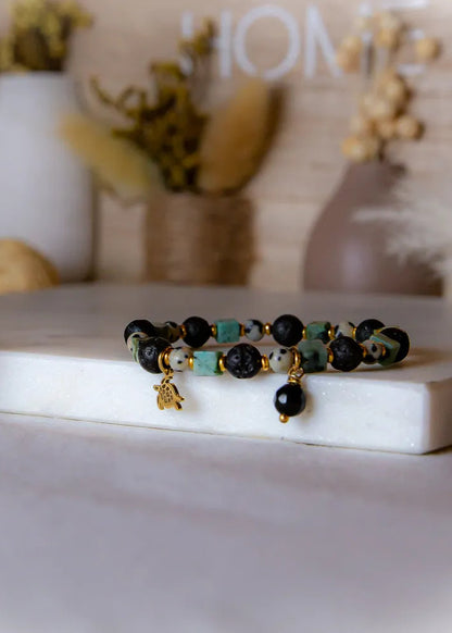 Afrykanska Harmonia Bransoletka Z Kamieniami - zdjęcie zbliżenia z koralików z lawy, kamienia dalmatyńskiego, zielonych kostek, złotego żółwia i czarnej kropli, na białym tle z elementami dekoracyjnymi.