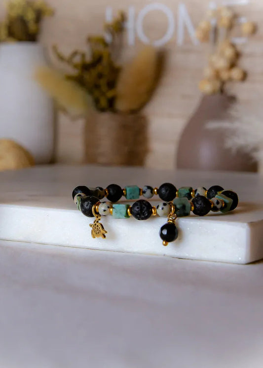 Afrykanska Harmonia Bransoletka Z Kamieniami - zdjęcie zbliżenia z koralików z lawy, kamienia dalmatyńskiego, zielonych kostek, złotego żółwia i czarnej kropli, na białym tle z elementami dekoracyjnymi.
