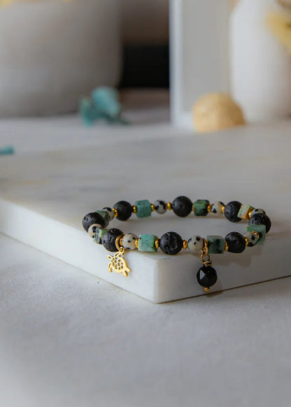 Afrykańska Harmonia Bransoletka Z Kamieniami z turkusowymi i czarnymi kamieniami, złotym żółwikiem i zawieszką na marmurowej podstawie.