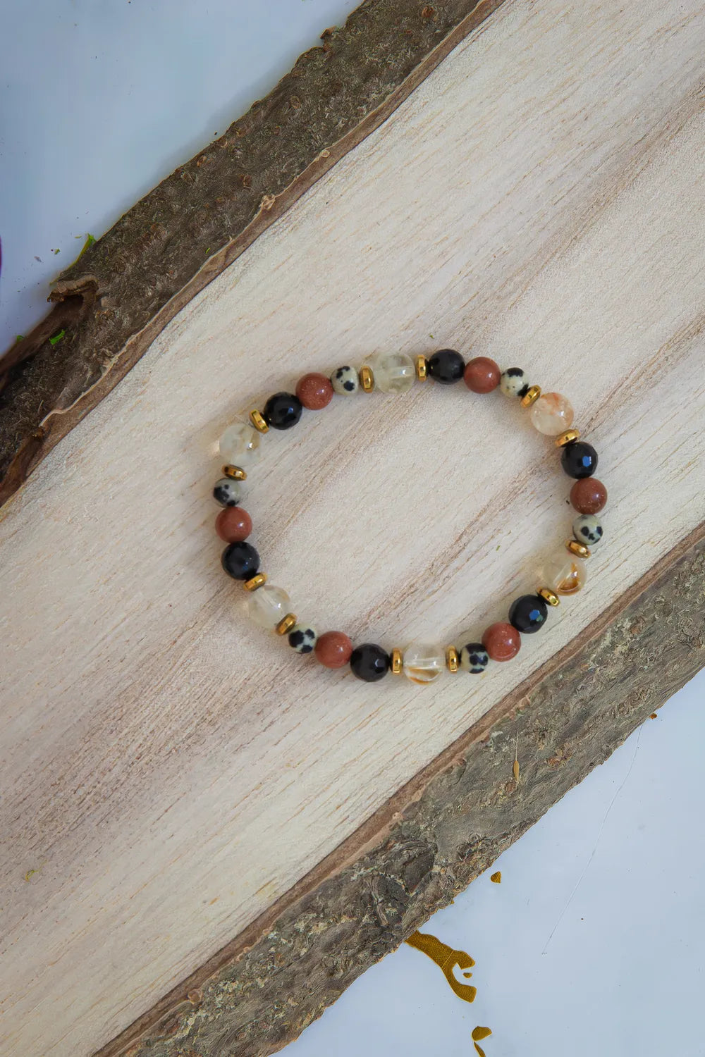 ALCHEMIA SUKCESU - BRANSOLETKA Z CYTRYNU, PIASKU PUSTYNI I KRYSZTAŁU GÓRSKIEGO. Bransoletka z kolorowymi koralikami, w tym cytrynami, piaskiem pustyni i kryształem górskim, ułożona na drewnianym tle.