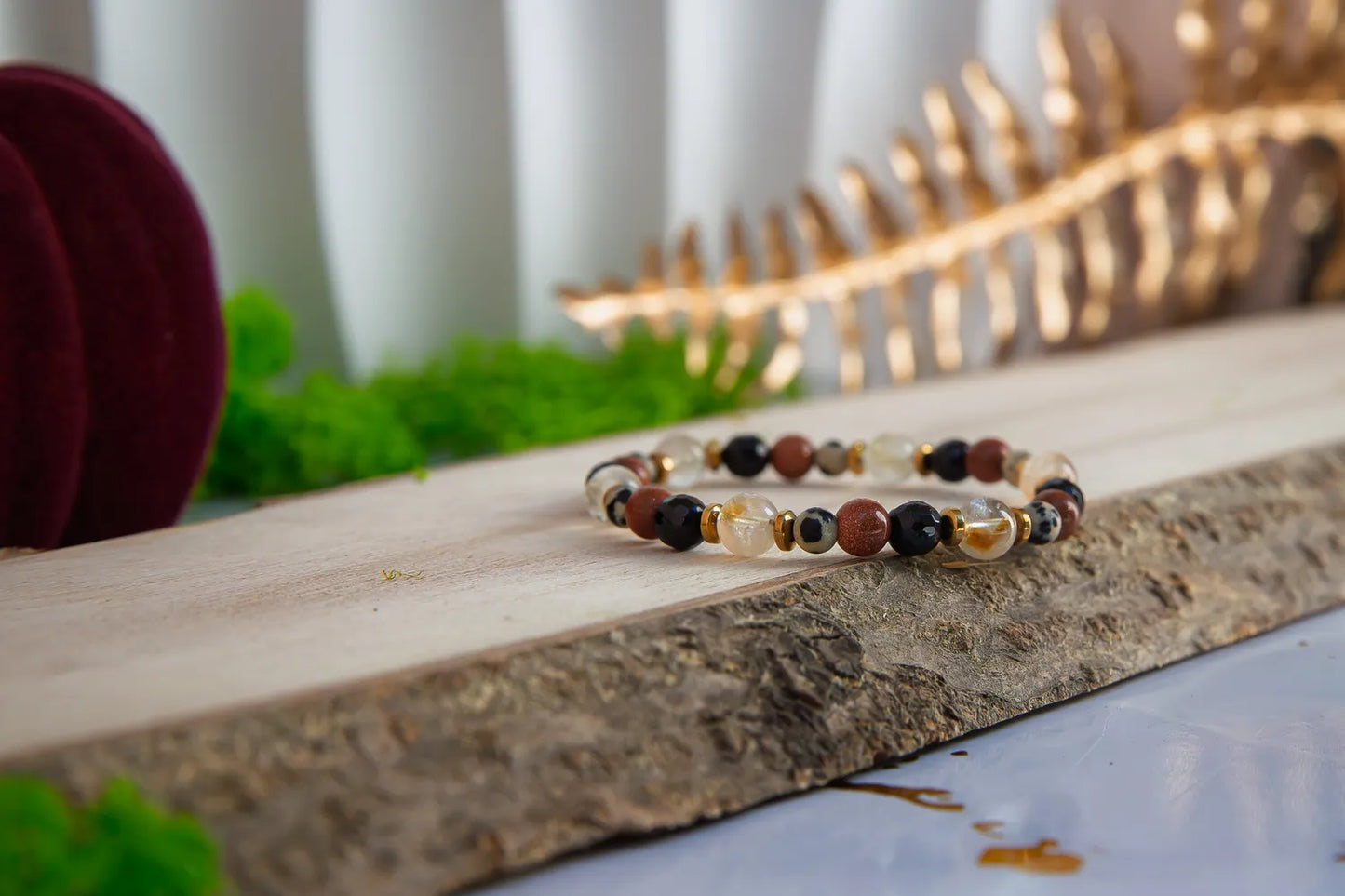 ALCHEMIA SUKCESU - BRANSOLETKA Z CYTRYNU, PIASKU PUSTYNI I KRYSZTAŁU GÓRSKIEGO. Bransoletka z kolorowymi koralikami, w tym cytrynami, piaskiem pustyni i kryształem górskim, leży na drewnianym pomoście z zielonym tłem i złotym elementem w tle.