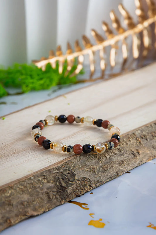 ALCHEMIA SUKCESU - BRANSOLETKA Z CYTRYNU, PIASKU PUSTYNI I KRYSZTAŁU GÓRSKIEGO. Bransoletka z kolorowymi koralikami, w tym cytrynami, piaskiem pustyni i kryształem górskim, leży na drewnianym tle z zielonymi roślinami w tle.