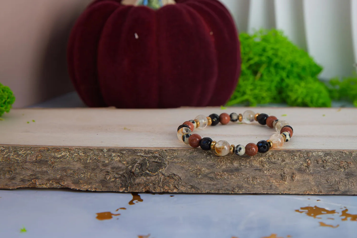 ALCHEMIA SUKCESU - BRANSOLETKA Z CYTRYNU, PIASKU PUSTYNI I KRYSZTAŁU GÓRSKIEGO. Bransoletka z kolorowymi koralikami leży na drewnianym bloku, na tle czerwonej dyni i zielonych roślin.