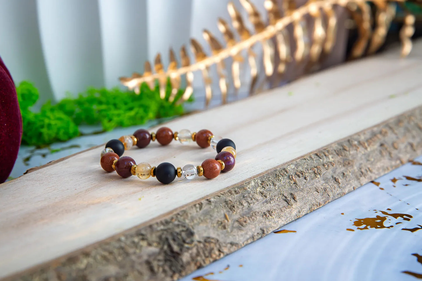 AMULET KRÓLOWEJ - BRANSOLETKA Z ONYKSU, CYTRYNU I JASPISU MORSKIEGO. Bransoletka z kolorowymi koralikami z onyksu, cytrynu i jaspisu morskiego leży na drewnianym tle z zielonymi roślinami i złotą gałązką w tle.