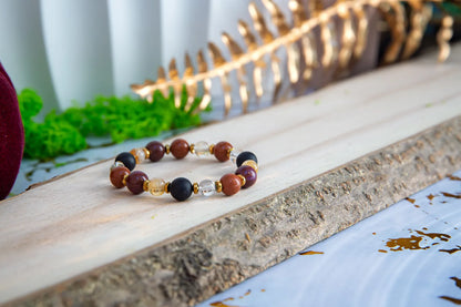 AMULET KRÓLOWEJ - BRANSOLETKA Z ONYKSU, CYTRYNU I JASPISU MORSKIEGO. Bransoletka z kolorowymi koralikami z onyksu, cytrynu i jaspisu morskiego leży na drewnianym tle z zielonymi roślinami i złotą gałązką w tle.