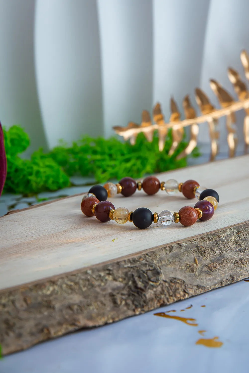 AMULET KRÓLOWEJ - BRANSOLETKA Z ONYKSU, CYTRYNU I JASPISU MORSKIEGO. Bransoletka z kolorowymi kamieniami na drewnianym tle, otoczona zielenią i złotą dekoracją.
