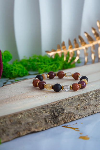 AMULET KRÓLOWEJ - BRANSOLETKA Z ONYKSU, CYTRYNU I JASPISU MORSKIEGO. Bransoletka z kolorowymi kamieniami na drewnianym tle, otoczona zielenią i złotą dekoracją.