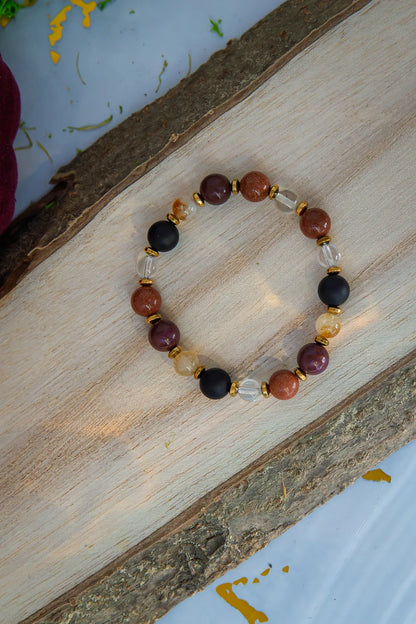 AMULET KRÓLOWEJ - BRANSOLETKA Z ONYKSU, CYTRYNU I JASPISU MORSKIEGO. Bransoletka z kolorowymi kamieniami, w tym czarnym onyksu, pomarańczowym cytrynu i jaspisu morskiego, ułożona na drewnianym tle.