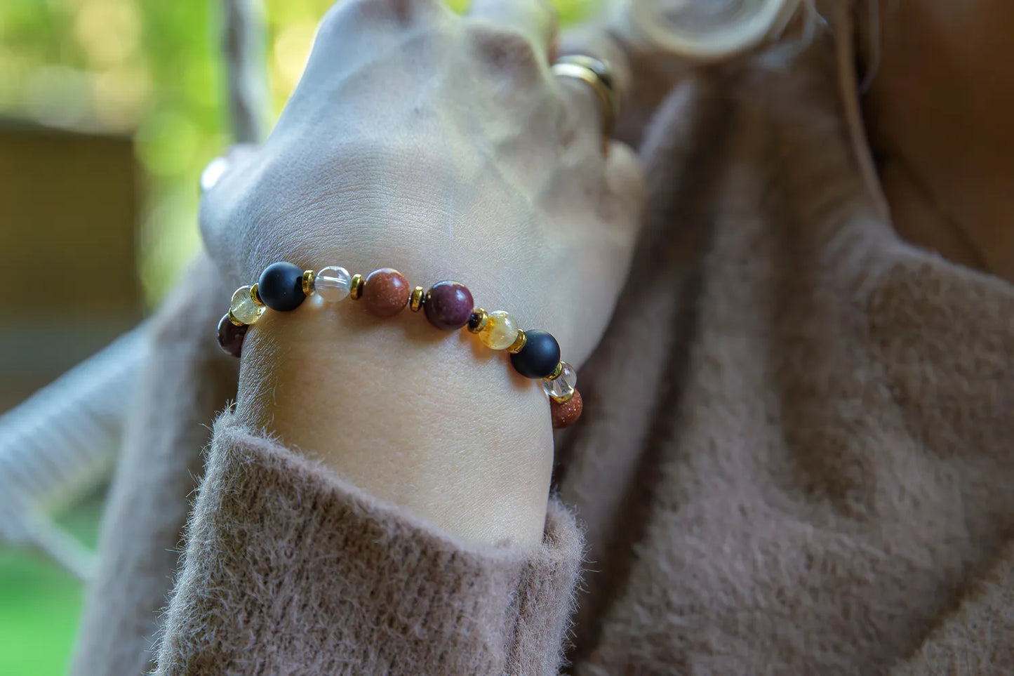 AMULET KRÓLOWEJ - BRANSOLETKA Z ONYKSU, CYTRYNU I JASPISU MORSKIEGO. Bransoletka z kolorowymi kamieniami, w tym czarnym onyksu, żółtym cytrynu i różowym jaspisu morskiego, na tle delikatnej, beżowej tkaniny.