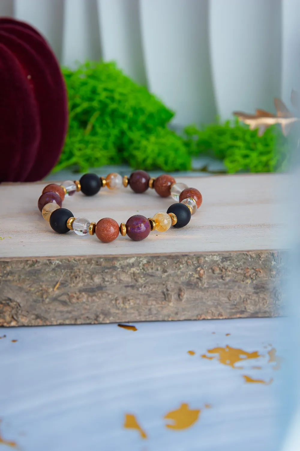AMULET KRÓLOWEJ - BRANSOLETKA Z ONYKSU, CYTRYNU I JASPISU MORSKIEGO. Bransoletka z kolorowymi koralikami z onyksu, cytrynu i jaspisu morskiego, ułożona na drewnianym bloku z zielonym mchem w tle.