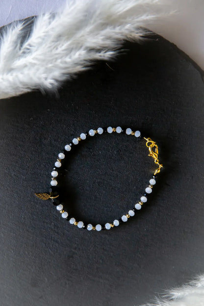 Anielskie Skrzydlo Bransoletka Z Turmalinami I Hematytami: złotą zawieszkę skrzydła, czarne i jasno-niebieskie kamienie, złotymi kuleczkami, na ciemnym tle z piórkami.