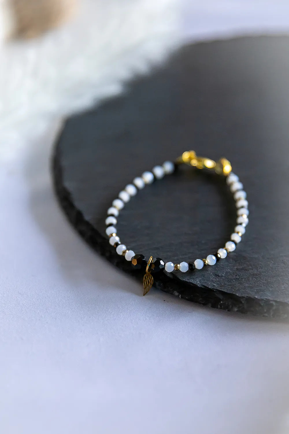 Anielskie Skrzydlo Bransoletka Z Turmalinem I Hematytem - subtelna bransoletka z białych i czarnych kamieni szlachetnych, ze złotym zapięciem i zawieszką skrzydło, na ciemnej powierzchni.