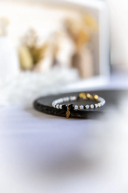 Anielskie Skrzydło Bransoletka Z Turmalinu I Hematytu: czarny turmalin i białe kamienie z złotym zawieszeniem skrzydła na ciemnej podstawie.