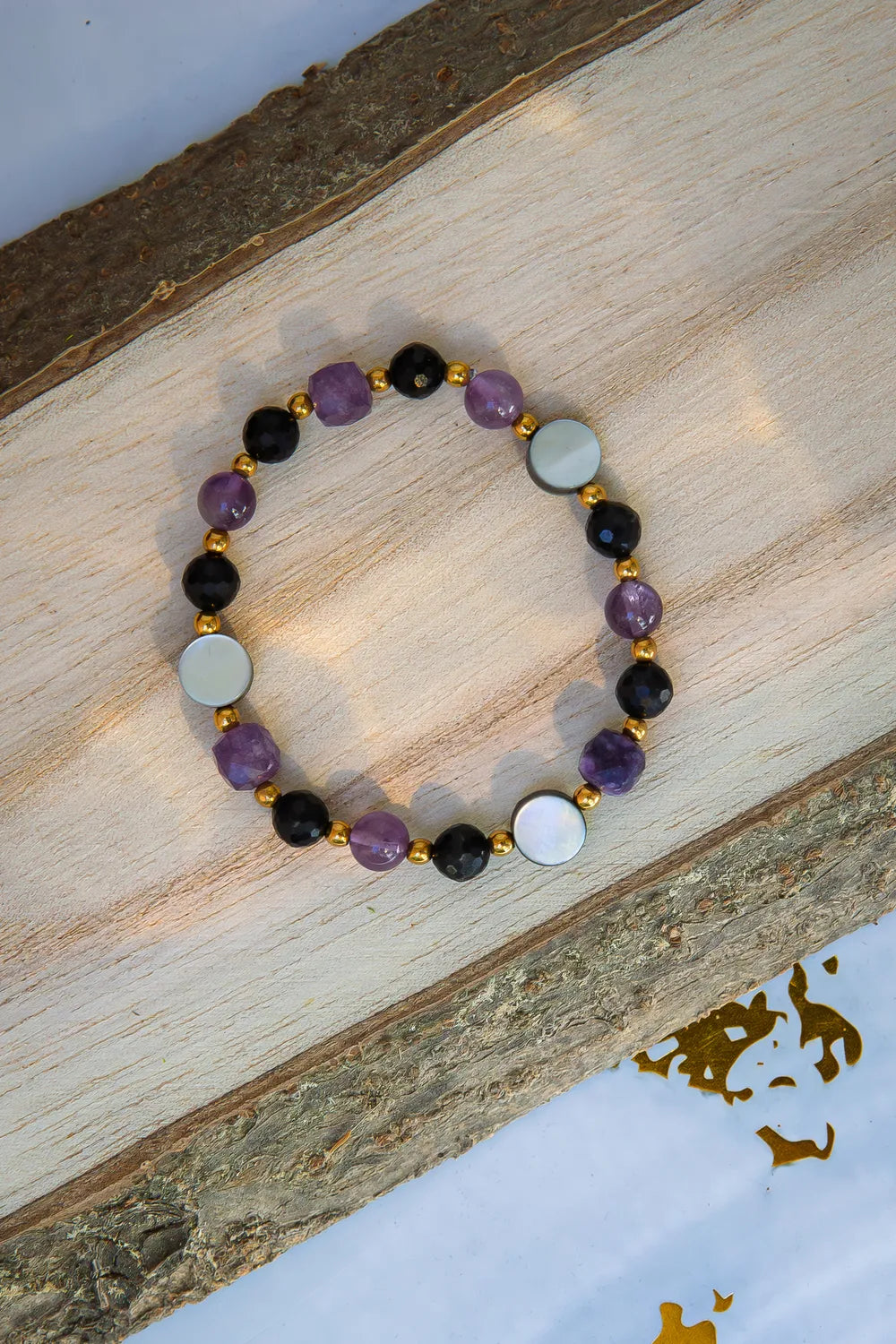 BLASK KSIĘŻYCA - BRANSOLETKA Z AMETYSTU, MASY PERŁOWEJ I CZARNEGO TURMALINU. Bransoletka z kolorowymi kamieniami, w tym ametystem, masą perłową i czarnym turmalinem, ułożona na drewnianym tle.