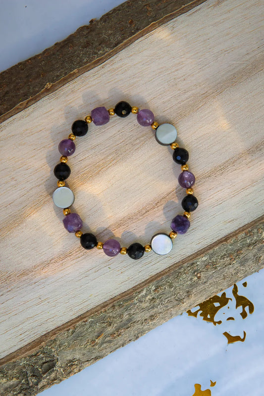BLASK KSIĘŻYCA - BRANSOLETKA Z AMETYSTU, MASY PERŁOWEJ I CZARNEGO TURMALINU. Bransoletka z kolorowymi kamieniami, w tym ametystem, masą perłową i czarnym turmalinem, ułożona na drewnianym tle.
