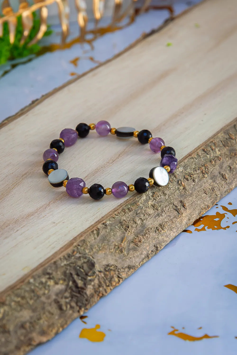 BLASK KSIĘŻYCA - BRANSOLETKA Z AMETYSTU, MASY PERŁOWEJ I CZARNEGO TURMALINU. Bransoletka z ametystem, czarnym turmalinem i masą perłową leży na drewnianym bloku z zielonym mchem i złotymi liśćmi w tle.