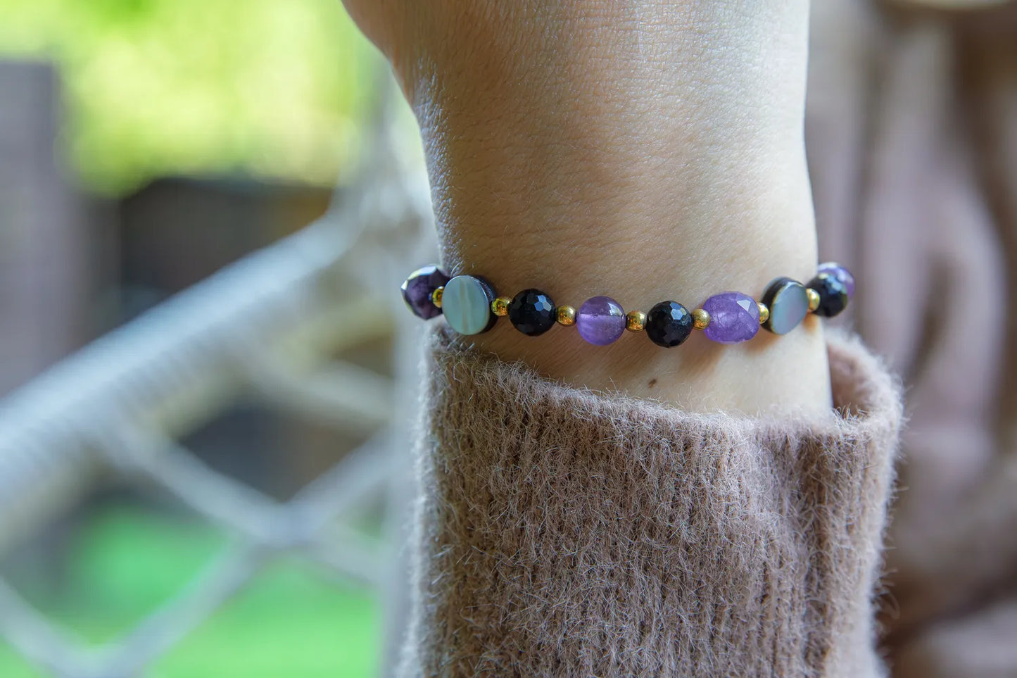 BLASK KSIĘŻYCA - BRANSOLETKA Z AMETYSTU, MASY PERŁOWEJ I CZARNEGO TURMALINU. Bransoletka z kolorowymi kamieniami, w tym ametystem, masą perłową i czarnym turmalinem, na tle delikatnej bluzki. Ułożenie na nadgarstku.