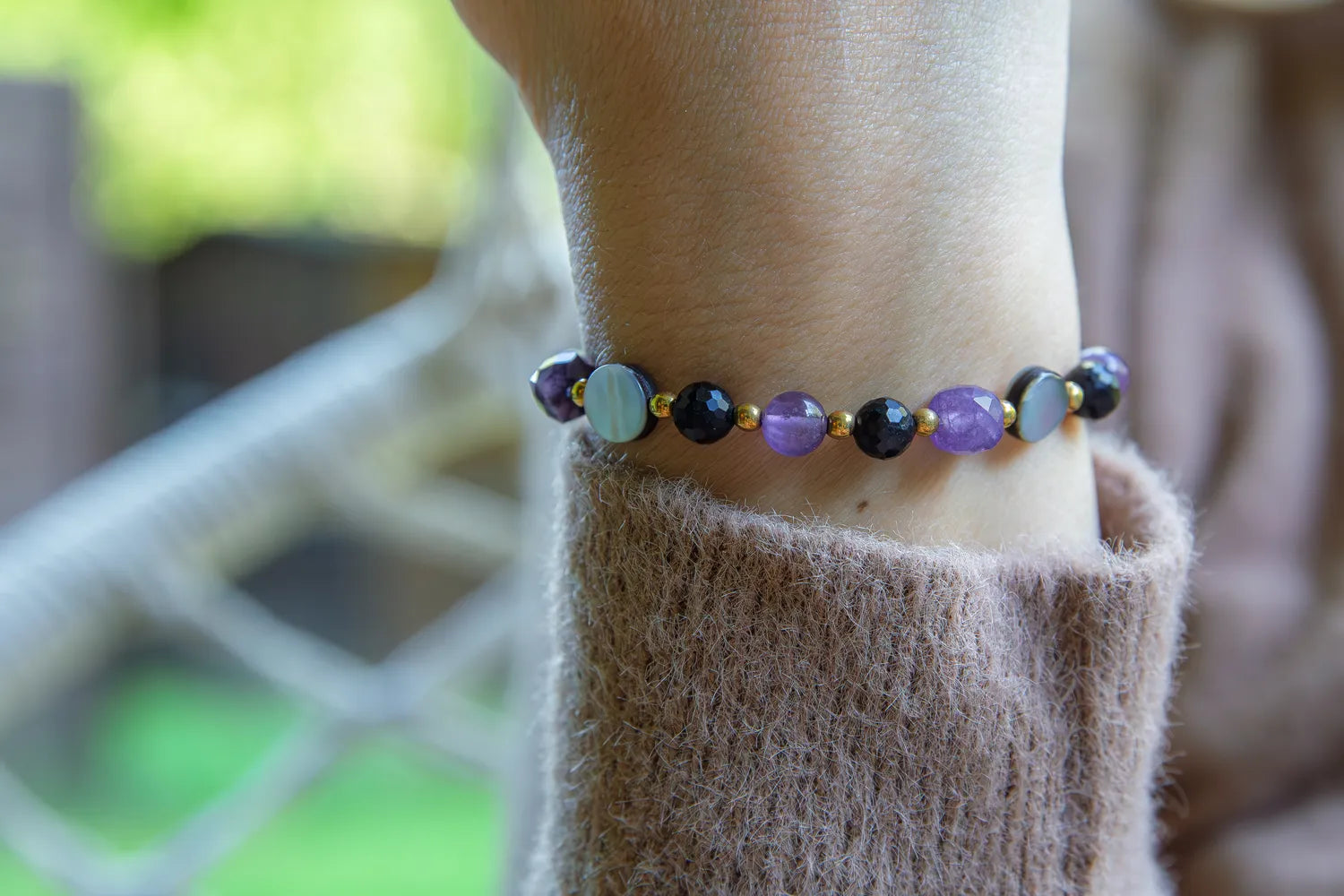 BLASK KSIĘŻYCA - BRANSOLETKA Z AMETYSTU, MASY PERŁOWEJ I CZARNEGO TURMALINU. Bransoletka z kolorowymi kamieniami, w tym ametystem, masą perłową i czarnym turmalinem, na tle delikatnej bluzki. Ułożenie na nadgarstku.