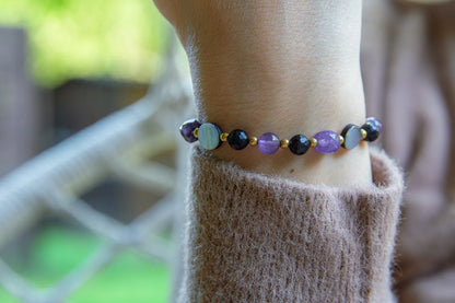 BLASK KSIĘŻYCA - BRANSOLETKA Z AMETYSTU, MASY PERŁOWEJ I CZARNEGO TURMALINU. Bransoletka z kolorowymi kamieniami, w tym ametystem, masą perłową i czarnym turmalinem, na tle delikatnej bluzki. Ułożenie na nadgarstku.