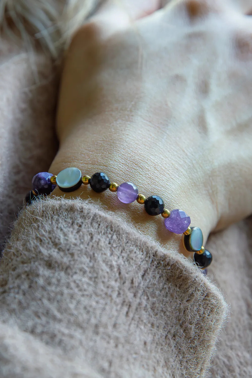 BLASK KSIĘŻYCA - BRANSOLETKA Z AMETYSTU, MASY PERŁOWEJ I CZARNEGO TURMALINU. Bransoletka z kolorowymi kamieniami, w tym ametystem, masą perłową i czarnym turmalinem, na tle delikatnej, beżowej tkaniny.