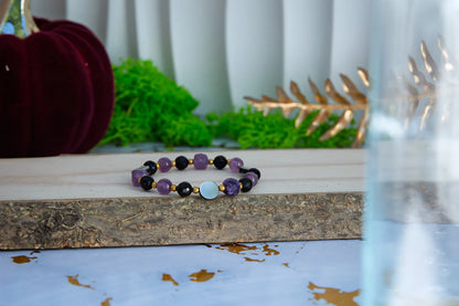 BLASK KSIĘŻYCA - BRANSOLETKA Z AMETYSTU, MASY PERŁOWEJ I CZARNEGO TURMALINU. Bransoletka z ametystem, czarnym turmalinem i masą perłową leży na drewnianym bloku z zielonym mchem i złotymi liśćmi w tle.