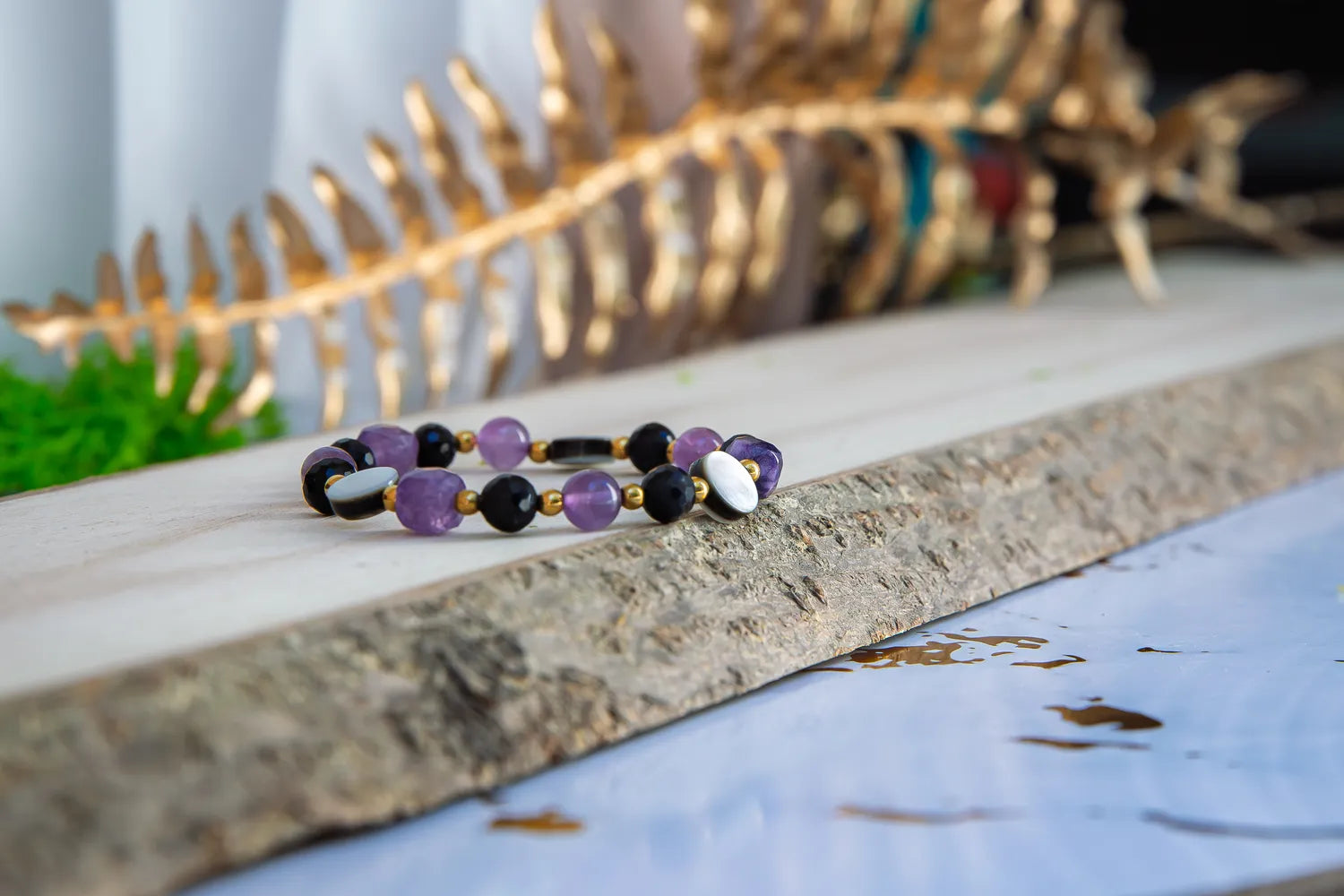 BLASK KSIĘŻYCA - BRANSOLETKA Z AMETYSTU, MASY PERŁOWEJ I CZARNEGO TURMALINU. Bransoletka z kolorowymi kamieniami, w tym ametystem, masą perłową i czarnym turmalinem, leży na drewnianym tle z złotym liściem w tle.
