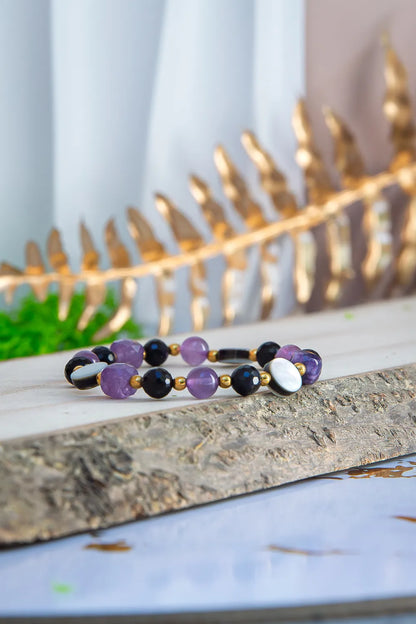 BLASK KSIĘŻYCA - BRANSOLETKA Z AMETYSTU, MASY PERŁOWEJ I CZARNEGO TURMALINU. Bransoletka z kolorowymi kamieniami, w tym ametystem, czarnym turmalinem i masą perłową, ułożona na kamieniu na tle złotej liściny.