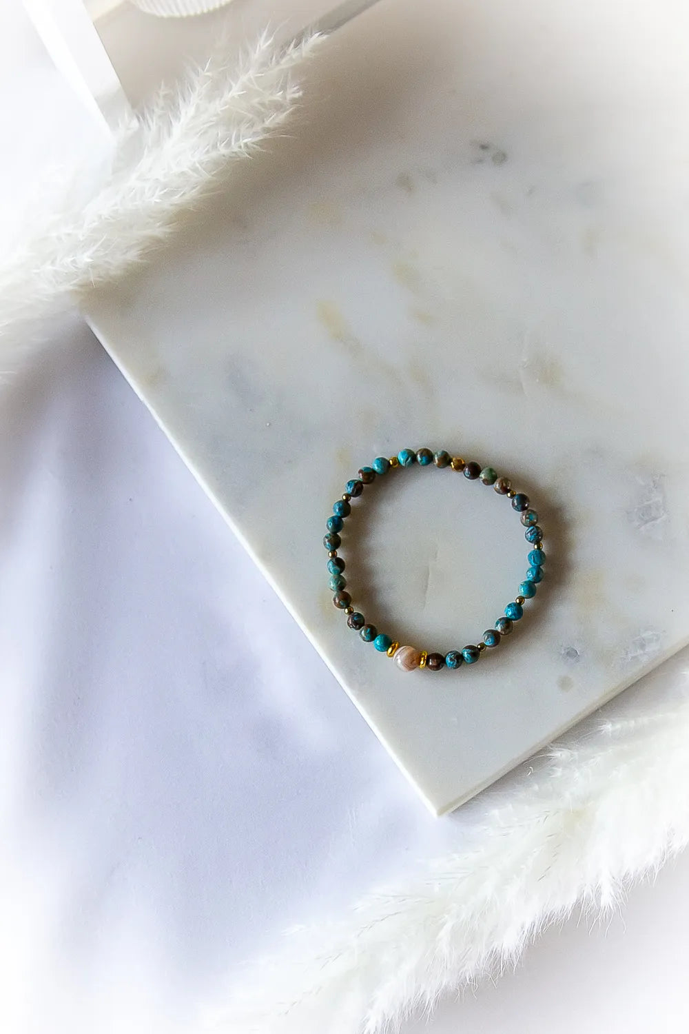 Błękitna Równowaga Bransoletka Agat Turkus z plecionymi kamieniami w odcieniach błękitu oraz złotymi przekładkami na marmurowej płytce.